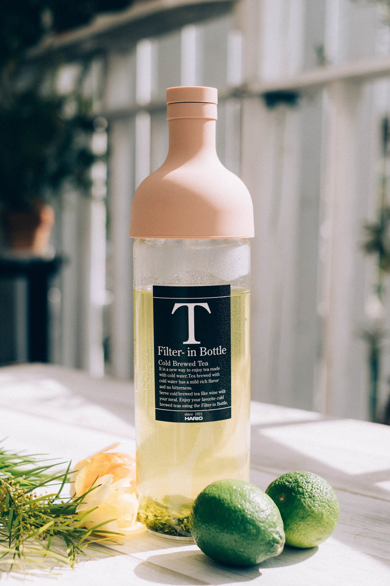 HARIO Filter-in Bottle 750ml smoky pink filled with cold-brewed tea, with limes and a flower on a white table
