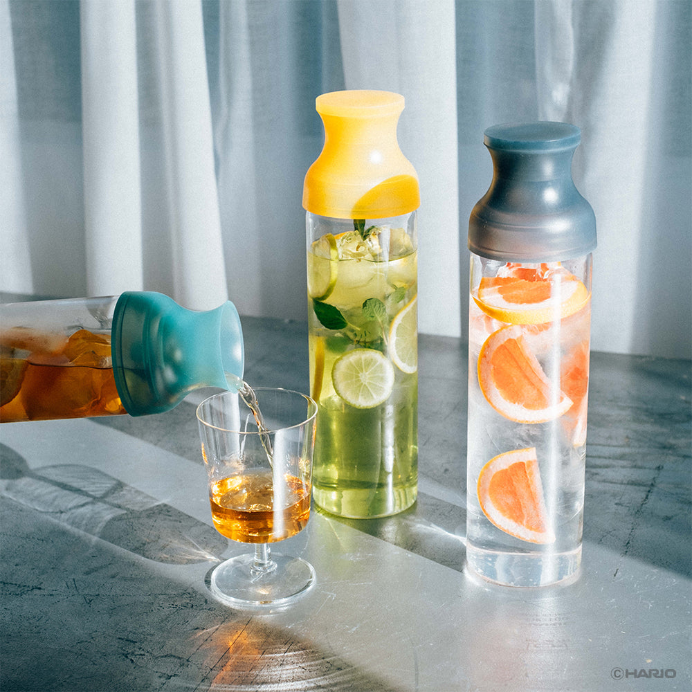 Pouring cold-brewed tea from a HARIO Filter-in Carafe with a green cap into a glass, beside yellow and grey carafes