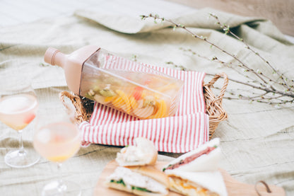 HARIO Ka-Ku Bottle 1,200ml smoky pink filled with sliced fruit, lying in a picnic basket with glasses and sandwiches