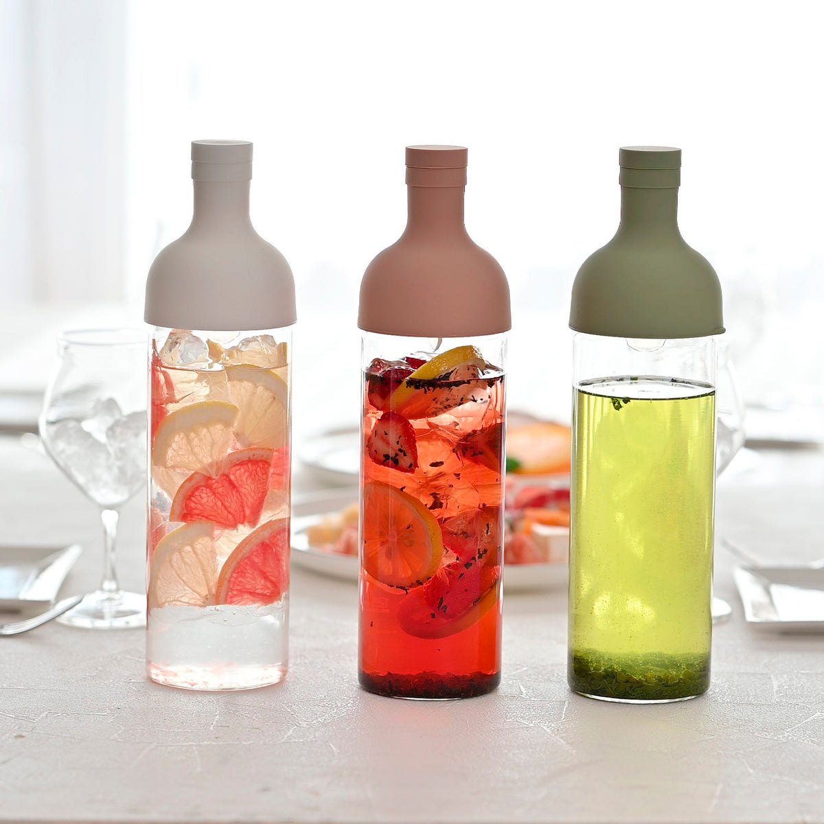Three HARIO Filter-in Bottles 750ml in white, smoky pink and smoky green on a table, filled with fruit teas and cold-brewed sencha