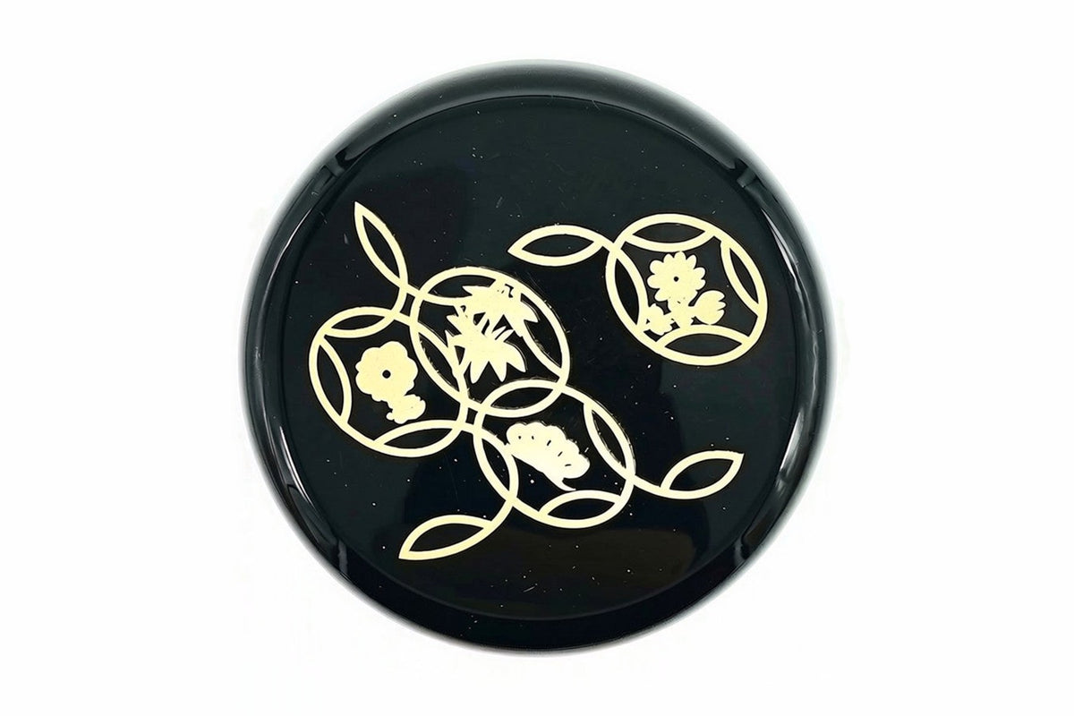 Lid of the black Hana-Shippō Natsume from above, gold circles holding a plum blossom, bamboo leaves, pine and a small flower