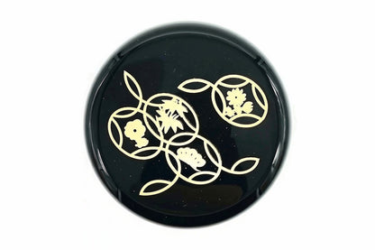 Lid of the black Hana-Shippō Natsume from above, gold circles holding a plum blossom, bamboo leaves, pine and a small flower