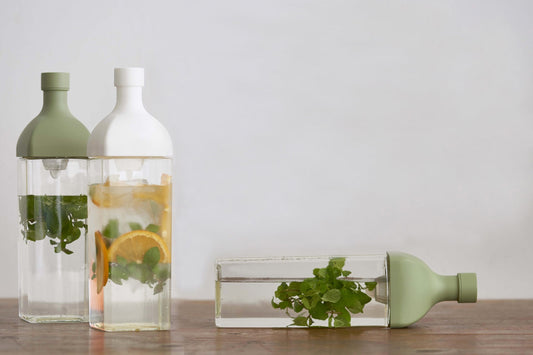 HARIO Ka-Ku Bottles in smoky green and white standing on a wooden table, with a third lying on its side, all filled with mint and citrus cold brew