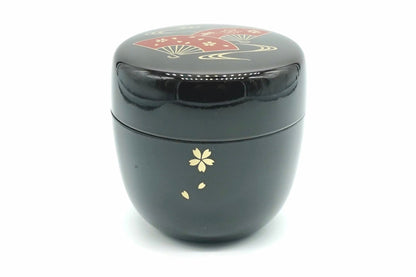 Ōgi Natsume in black, a glossy matcha caddy with a gold cherry blossom and falling petals on the body and red folding fans on the lid