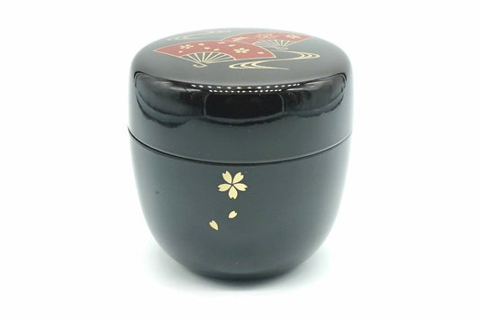 Ōgi Natsume in black, a glossy matcha caddy with a gold cherry blossom and falling petals on the body and red folding fans on the lid