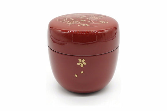 Ōgi Natsume in red, a glossy matcha caddy with a gold cherry blossom and falling petals on the body and folding fans on the lid