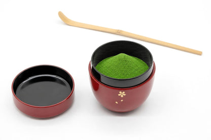 Ōgi Natsume in red, open, with matcha heaped inside, its lid set beside it with the inside facing up, and a bamboo chashaku behind