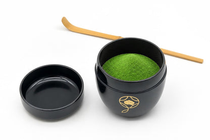 Black natsume with a gold shippō design, open, with matcha heaped inside, its lid set beside it with the inside facing up, and a bamboo chashaku behind